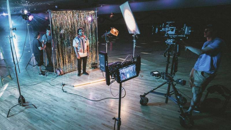 Videographer shooting talent with lighting set up and crew in background.