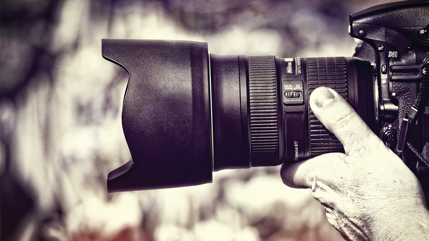  A close-up of a photographer's hand holding a camera lens with a lens hood attached.